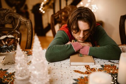 Person wearing a holiday sweater, sitting at a table with his head resting on his hands, gazing downward looking forlorn. Holiday decor is around.